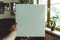 Hand holding blank white square paper cover album vinyl record wrap with plastic shrink texture against living room painting indoors plant.