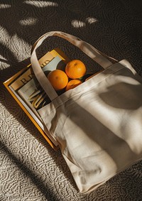 An open tote bag lying on the ground filled with book and two oranges accessories grapefruit accessory.