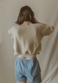 A woman in blue shorts and a beige oversized sweater is posing with her arms behind her back against a beige background clothing knitwear apparel.