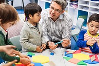 A male teacher, interacts with young children, Asian and Hispanic, in a classroom. The teacher and children are engaged in a classroom activity. Diverse kids at elementary school with teacher.