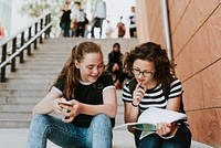 Two young girls sitting on stairs, studying together. The girls are focused on their studies, holding books and papers. The girls are engaged in learning. Teenage girls studying and doing homework.