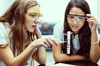 Two young girls, wearing safety goggles, in science experiment. The girls, engaged in science, use test tube. Safety goggles and science experiment. International students in experiment in classroom.