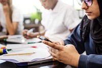 Asian female student in hijab using smartphone in class. Student focused on smartphone. Other students in background. Classroom setting with students and smartphones. Muslim teen student in classroom.