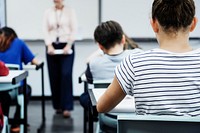 Students in a classroom, focused on their work. Classroom setting with students. Female teacher in the background. Classroom environment with students and teacher. High school students in classroom.