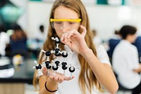 Young Caucasian girl in a science lab, holding a molecular model. The girl is focused on the molecular model, wearing safety goggles. Science lab, molecular model, girl. High school education.