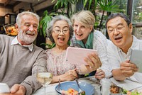 A diverse group of senior friends, both men and women, enjoying a meal together. Elderly friends taking selfies, laughing, and sharing moments. Happy senior friends bonding, taking a selfie at lunch.