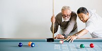 Two senior men, one Caucasian and one Asian, playing pool. The diverse senior men are focused on the pool game. Diverse senior man holds a cue stick, while the Asian man aims at the pool balls.