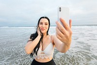 A woman taking a selfie at the beach. The woman smiles while holding her phone. Woman's relaxed and happy mood. Body positivity and inclusivity influencer taking selfie. Curvy latina woman at beach.