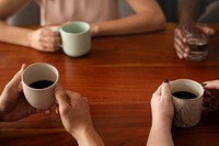 Diverse friends taking a break holding coffee cups and a glass of water. Coffee cups, hands, and wooden table are visible. Coffee cups and hands are the focus. Friends having coffee together at table.