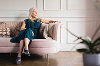 Elegant older woman wearing dress, sits on couch. The older woman looks thoughtful. Elegace old woman in luxury living room, calm old woman sitting thoughful, elderly woman in luxury vibes