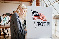 A woman voting at a polling station. The woman is casting vote. The voting booth has an American flag. The woman is focused on voting. Voting is taking place. Voter at American presidential election.