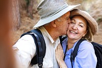 Elderly Caucasian couple hiking, man kissing woman on cheek. Happy elderly couple enjoying nature. Elderly couple with backpacks, smiling and hiking together. Senior couple enjoying outdoors adventure