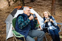 Elderly Caucasian couple camping, enjoy coffee. Happy elderly couple sit and smile in outdoors camping chairs with coffee mugs. Elderly Caucasian couple drink coffee, happy outdoor nature camping