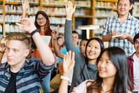 Diverse group of students in a classroom, raising hands to participate. Male and female students, various ethnicities, engaged in learning. Classroom, students, participation. High school education.