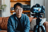 A camera recording video of an asian man sitting on a couch glasses tripod adult.