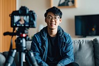 A camera recording video of an asian man sitting on a couch portrait tripod photo.