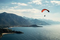 Paraglider flying over the sea with blue water and mountains paragliding recreation adventure. AI generated Image by rawpixel.