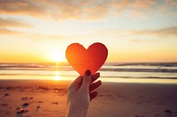 Hand holding a cut out of heart shaped paper against a sunset beach sunlight sky tranquility.