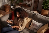 Young Black woman managing online banking with smartphone sitting on the sofa at home furniture reading adult.