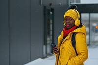 Happy young black woman holding mobile phone enjoying music listening through wireless headphones on footpath jacket adult coat.