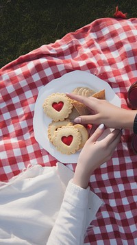 Hands reaching for heart-shaped cookies on a picnic blanket. Cookies on a plate, picnic setup, and hands reaching for treats. Outdoor picnic scene with cookies.