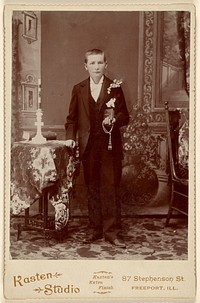 Unidentified young man standing, holding book and beads by Kasten Studio