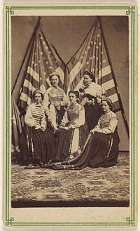 Five women, two standing, three seated, in costume, posed in front of two American flags