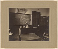 Portrait of a male mathematics teacher with moustache seated at his classroom desk