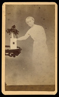 Female "spirit" standing next to a table with a photograph propped against a vase with flowers by William H Mumler and Helen F Stuart