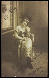 A man in drag (Charlie) sits face-on, wearing detailed costume, with a window painted on the backdrop. Photographic postcard, 1919.