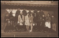 Soldiers, some in drag, pose on stage; two men in drag face each other in the centre. Photographic postcard, 191-.