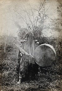 Minusinsk, Siberia: a Tartar medicine man or shaman in ceremonial dress with a covered face, holding a drum. Photograph, ca. 1920  of a photograph, 1900/1915.