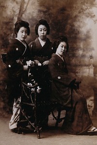 Three young women posing in a photographic studio, one seated on a bench, in front of a painted backdrop of a rural scene.