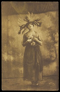 A performer in drag poses on stage wearing a head garment with sprawling feathers. Photographic postcard, 191-.