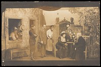 Actors, some in drag; with the attention focused on a character sitting at a table. Photographic postcard, 191-.