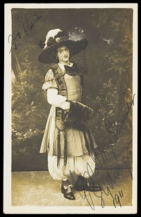 A man in drag wearing a very large hat with a veil, stands in front of a painted backdrop, with dense foliage. Photographic postcard, 1911.