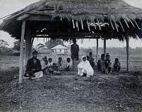 Assam, India: a kala-azar out-patient centre: a small group of men, women and children sit beneath a grass-roofed shelter with some metal bowls (for heating water for sterilisation ). Photograph, 1900/1920 .