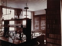 The interior of a pharmaceutical or chemical laboratory with a bench in the centre and books and papers on shelves around the walls. Photograph.