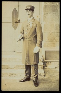 A man dressed wearing a long coat and a hat bearing the word "Cook", stands outside a building. Photographic postcard, 191-.