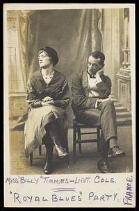 Two soldiers, one in drag called "Miss Billy Timmins", are seated in front of a painted backdrop. Photographic postcard, 1917.