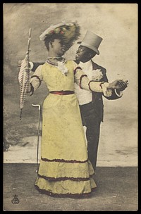 Two black actors, one in drag, dance together on stage. Coloured process print, ca. 1903.