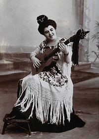 A seated woman, playing a guitar, wearing a fringed shawl. Photograph, ca.1900.