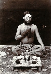 An Indian man wearing a Parsi string and make-up, squatting on a rug with his hand in a rosary bag, in a studio setting. Photograph, ca.1900.