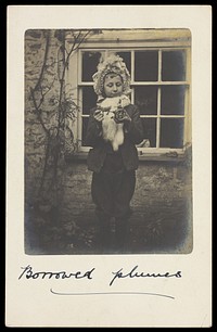 A boy dressed as a girl, wearing a bonnet; playing with a toy chariot. Photographic postcard, 1904.