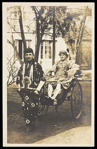 A man in ornate drag sits in a dog-cart and is pulled along the road by a man dressed in Chinese patterned robes and hat. Photographic postcard, 191-.