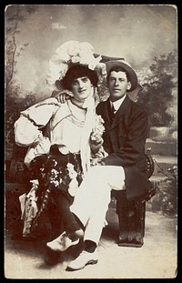 Two men, one in drag, sit together on a bench. Photographic postcard, 190-.
