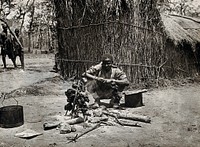 The Kilwa area (Nanganachi village ), Tanzania, East Africa: a Tanzanian man spit-roasting meat over an open fire in front of a grass hut. Photograph by Andrew Balfour, ca. 1910 .