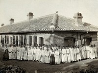 Leper asylum, Taiku, Korea: female patients wearing white robes stand in a group outside a brick building. Photograph, 1900/1920.