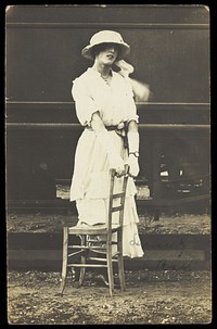 An soldier in a concert party poses in drag, wearing white and holding a chair, standing in front of a train. Photographic postcard, 191-.