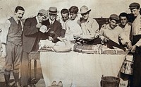 Staging of a leg amputation: the 'patient' lies on a table surrounded by men: one man poses with a saw, two men pretend to administer pain relief applying a teapot and a funnel to the patient's mouth. Photograph, 1905.
